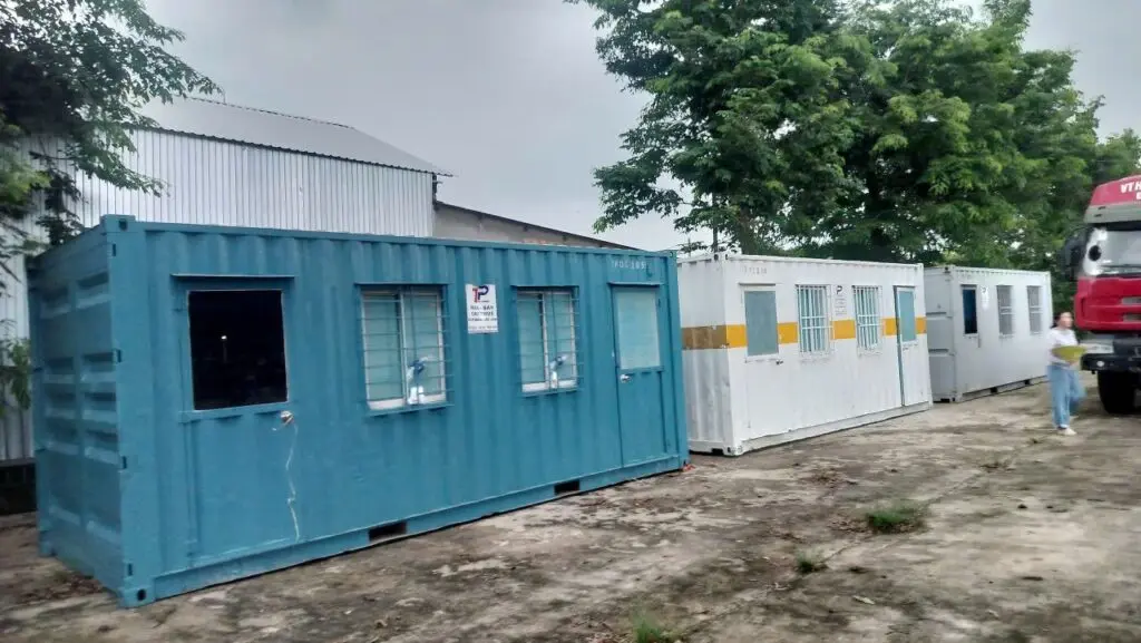 Container văn phòng sơn xanh ngọc cạnh container trắng vạch vàng và xe vận chuyển tại Bình Phước