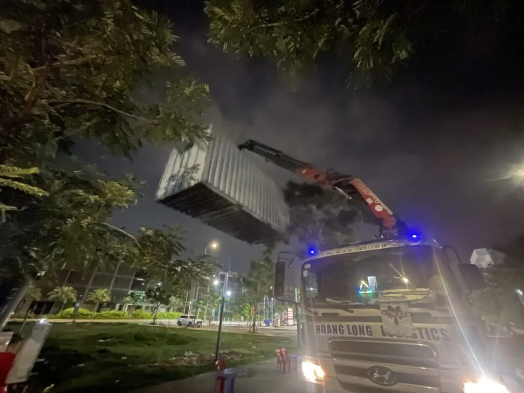 Xe cẩu nâng container văn phòng 20ft lên khỏi sàn xe tại Global City ban đêm